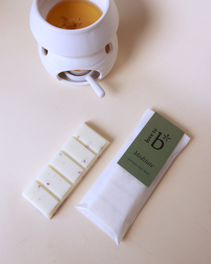A soy wax melt bar next to its packaging, with a white wax burner heating a melt in the background, all presented on a beige surface.