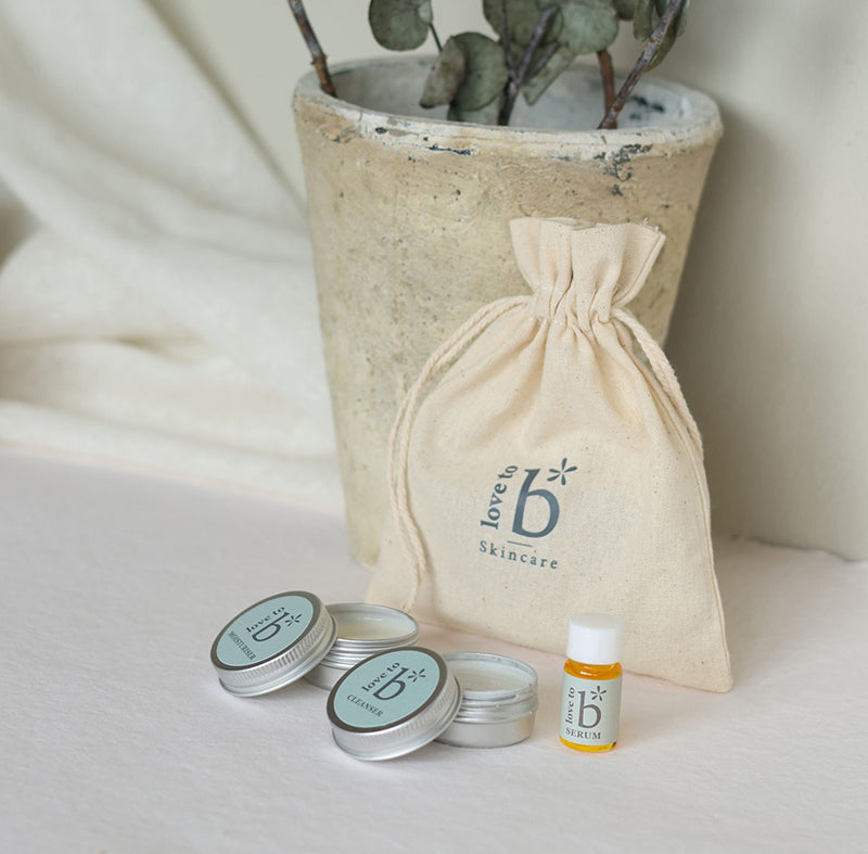 A natural skincare gift set featuring a beige drawstring bag with the 'love to b skincare' logo, three skincare tin products, and a small amber serum bottle, arranged in front of a rustic ceramic pot with eucalyptus branches on a draped cream fabric background.