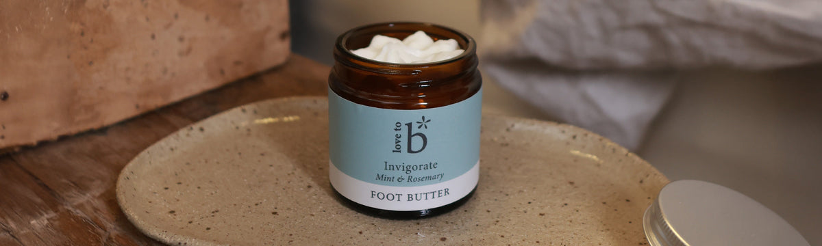 Love to b natural body butter and foot butter in signature scents on a plate next to a bed with the lid removed
