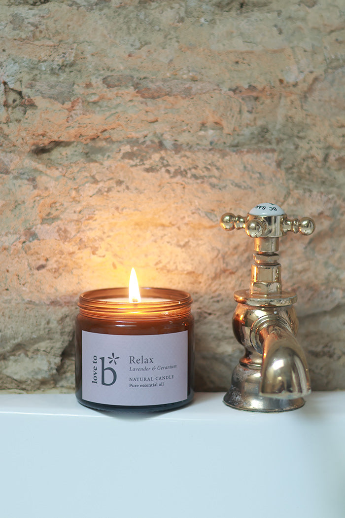 A lit natural soy candle sitting on a bath next to a tap. The label displays the scent - Lavender & Geranium.