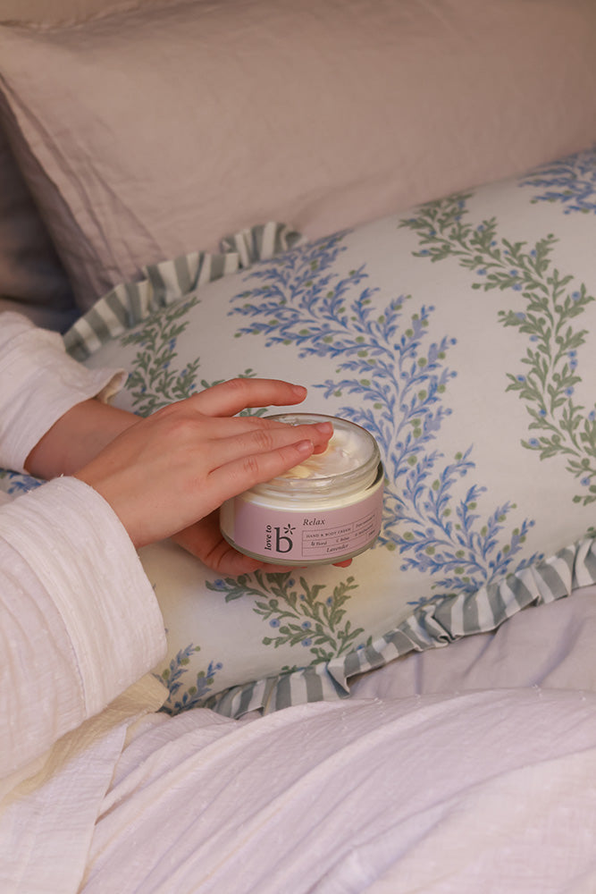 A person in a white robe holds an open jar of Love to b natural skincare cream over a patterned pillow in bed.