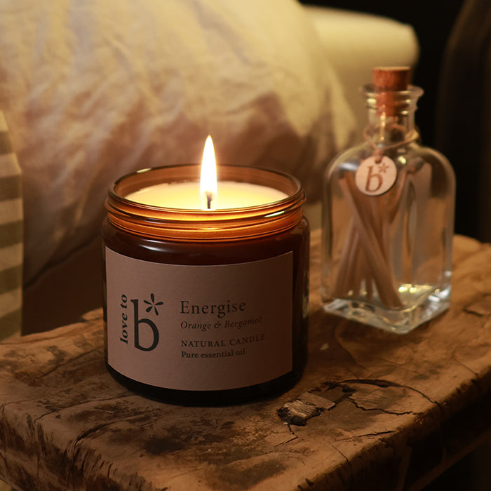A lit amber glass natural soy candle jar for Love to b, scented 'Energise Orange & Bergamot' sits on a rustic wooden surface beside a small clear glass bottle of matches with a cork stopper and a matching label, against a softly blurred indoor background.