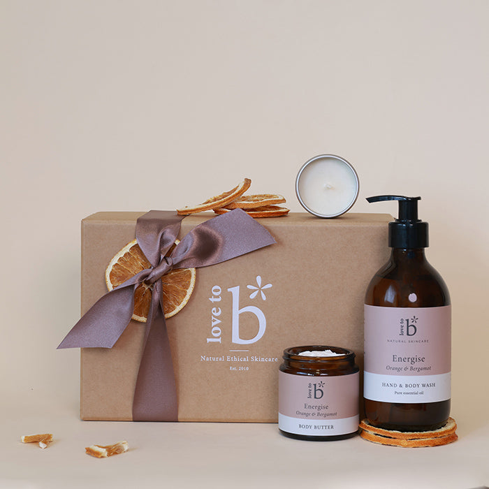 A curated natural wellness skincare gift set on a warm beige background, featuring a brown kraft gift box tied with a mauve ribbon and dried orange slices, alongside a white candle, a brown pump bottle of hand & body wash, and a small jar of body butter. The products display matching labels with the brand logo 'love to b'.
