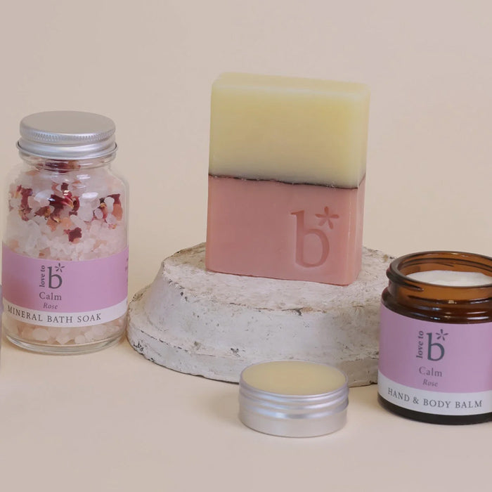 A set of natural skincare products from Love to b, including a natural soap bar, a glass jar of mineral bath soak, tin of natural lip balm, and an amber jar of hand and body balm, arranged on a neutral beige background with a rustic white pedestal.