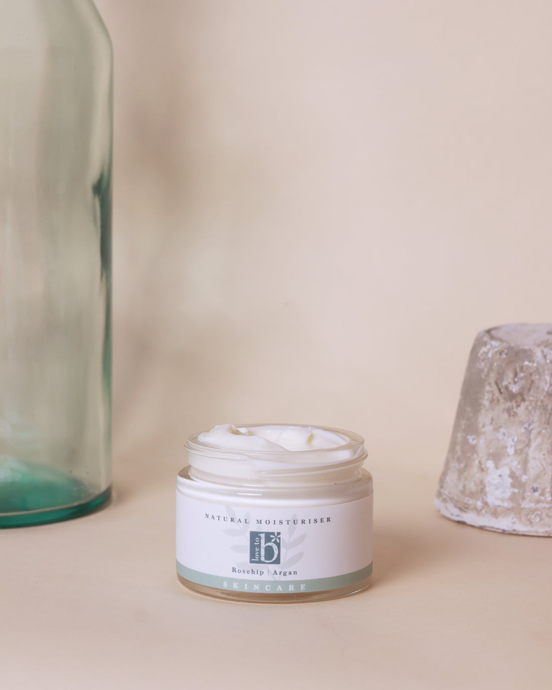 A natural facial moisturiser jar with a white label from Love to b, displayed next to a clear bottle and a textured bowl on a neutral background.