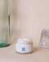 A natural facial moisturiser jar with a white label from Love to b, displayed next to a clear bottle and a textured bowl on a neutral background.