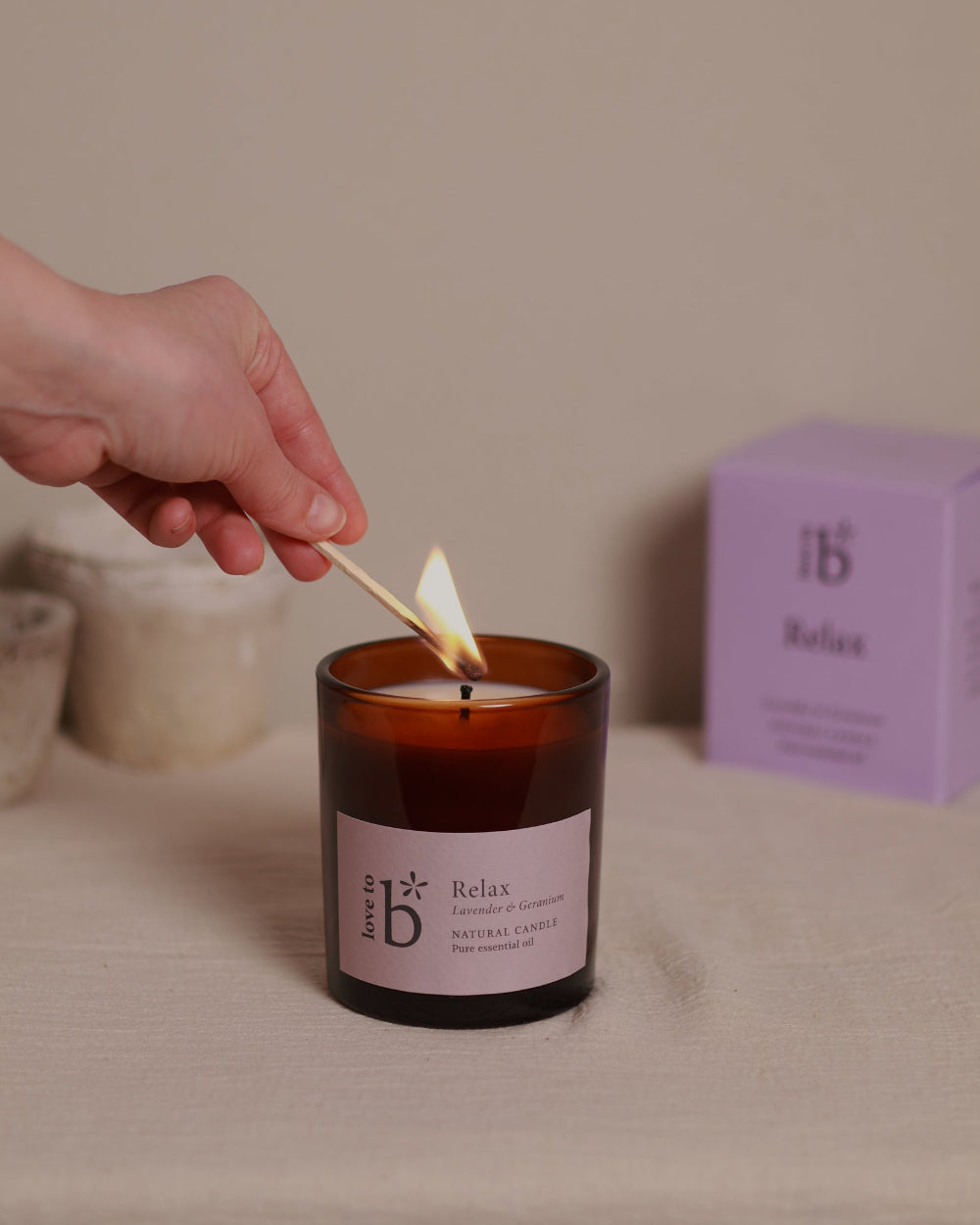 A lavender soy wax candle being lit in a clear glass jar with a label that reads &