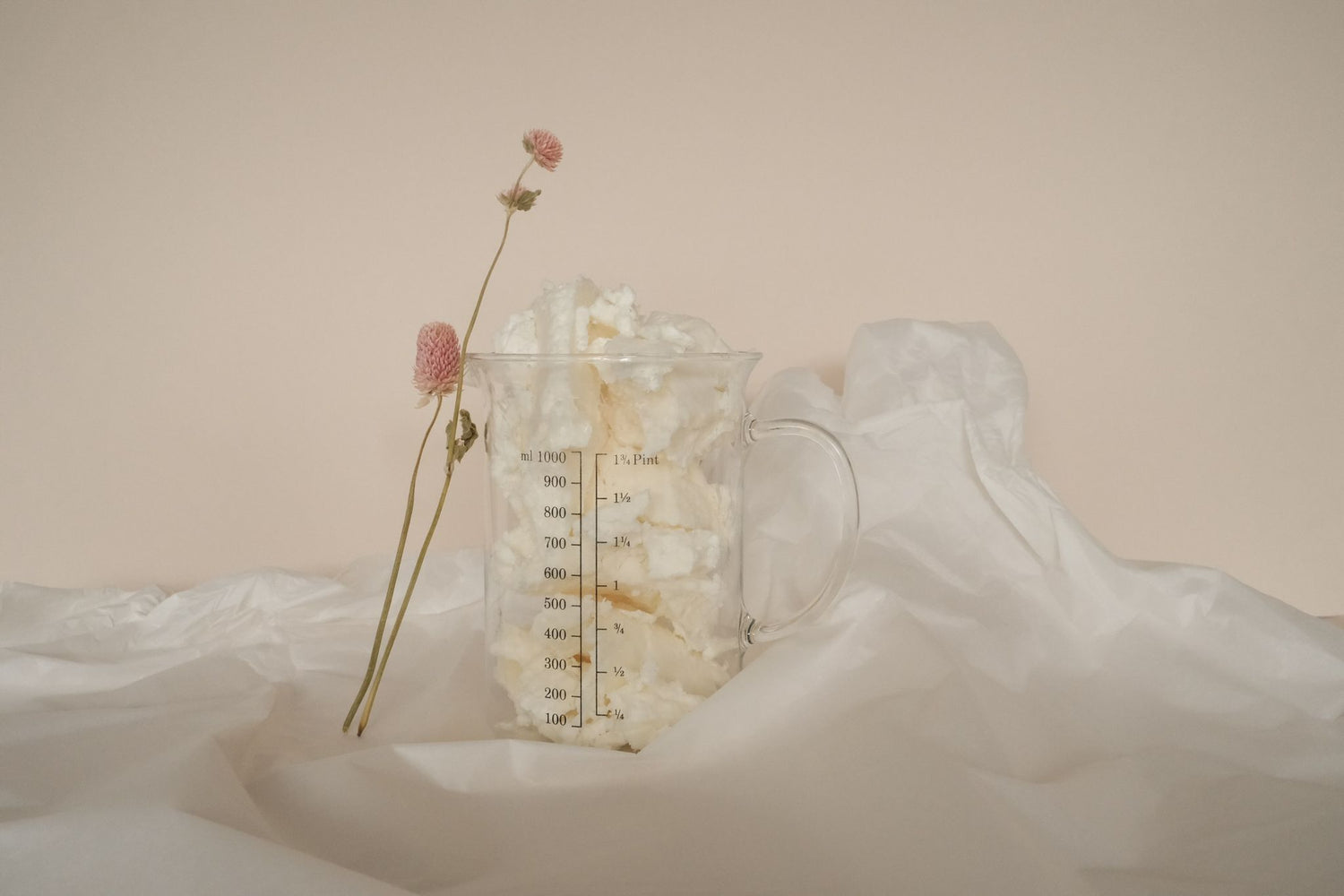 A glass beaker of Shea Butter with a pink flower leaning on it, set on a ruffled white sheet and a light peach coloured background.