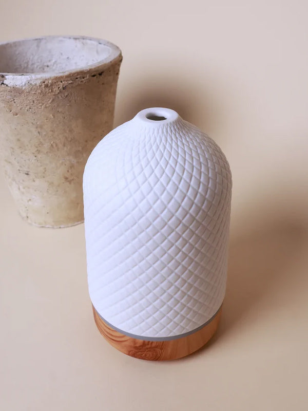 A white textured ceramic diffuser with a diamond-patterned body and a light wood base, shown on a beige background with a blurred concrete pot behind it.