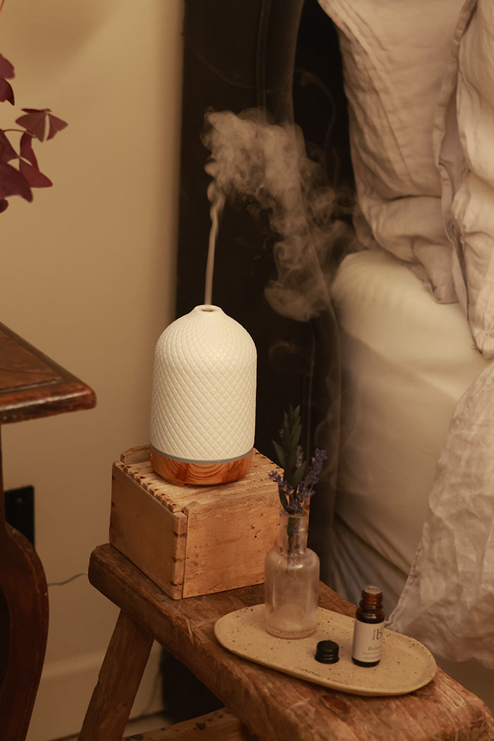 A white textured diffuser with a wood-toned base sits on a rustic wooden box on a bedside table, with mist rising from the top. A small amber glass bottle of essential oil blend is on a beige tray nearby, next to a bed with white bedding.
