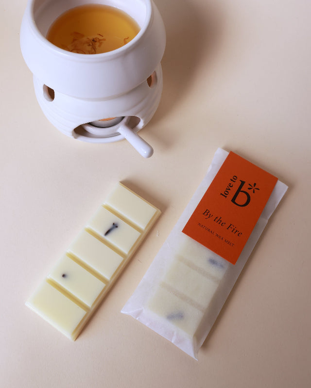 A wax burner with a lit wax melt in it, alongside a packaged natural wax melt bar labeled &