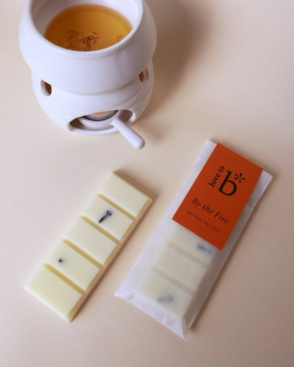 A wax burner with a lit wax melt in it, alongside a packaged natural wax melt bar labeled &