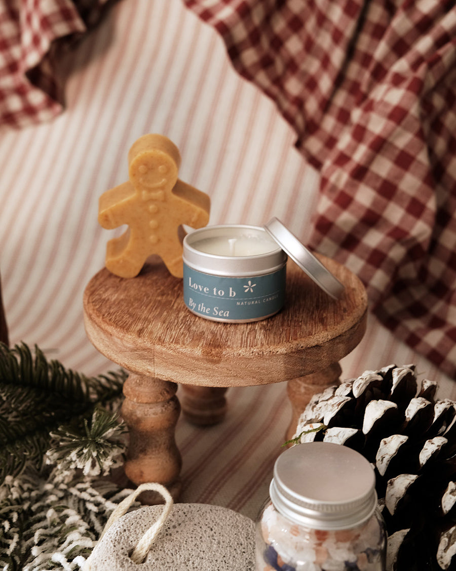 A candle with a silver lid sits on a round wooden stool, styled with a gingerbread man-shaped candle holder, pinecones, evergreen branches, and a red-and-white check blanket in the background.