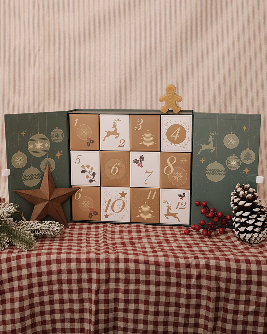 A Christmas advent calendar with 12 numbered square doors in beige, brown, and cream tones, displayed open on a red-and-white gingham tablecloth with holiday decorations.