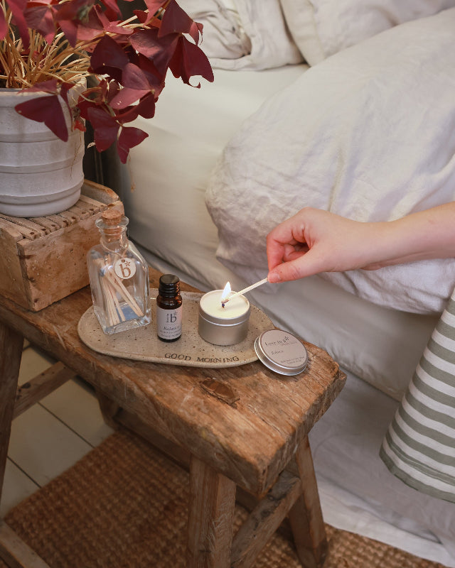 A natural soy candle tin being lit. Sat next to a amber bottle of essential oil in the balance scent. These products are next to a bed.
