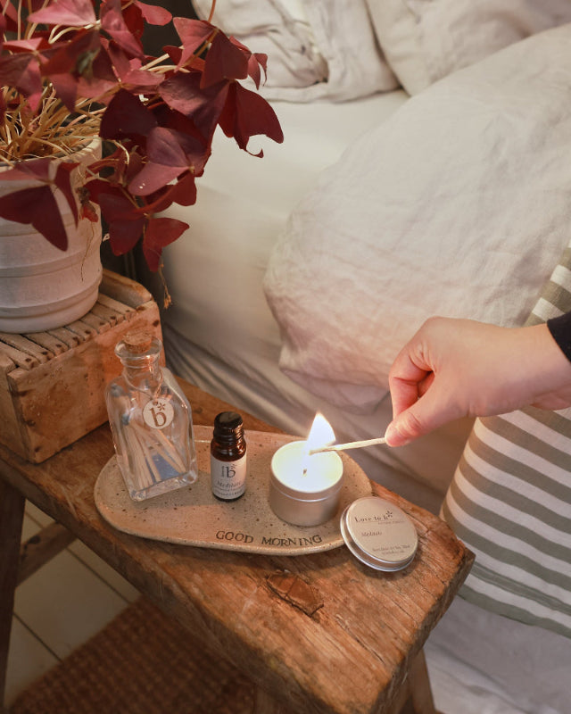 A small natural soy candle being lit. Sat next to a amber bottle of essential oil in the same scent. These products are next to a bed.