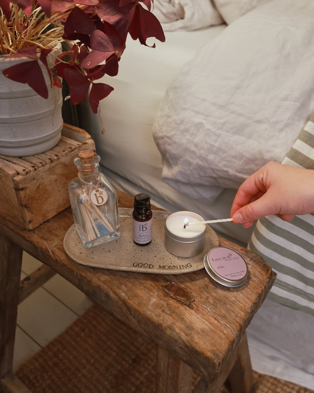 A natural soy candle being lit next to an amber bottle of essential oil in the same scent. These products are next to a bed.