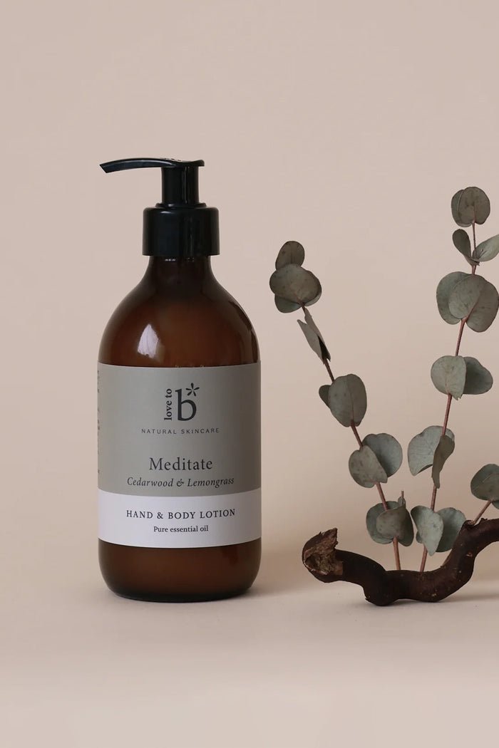 An amber glass pump bottle of Love to B Natural Skincare 'Meditate' hand and body lotion, styled next to a decorative eucalyptus branch on a warm beige background.