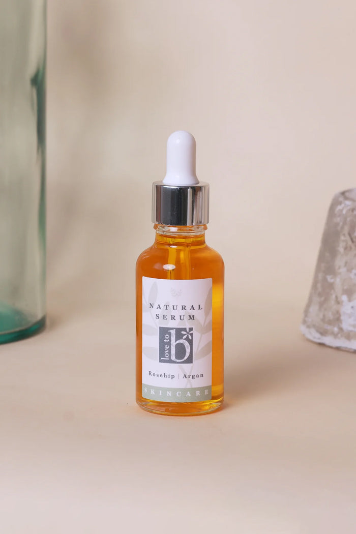 A bottle of Love to b Natural Serum with a white dropper cap and silver collar, containing rosehip and argan, set against a warm beige background with decorative glass props on either side.