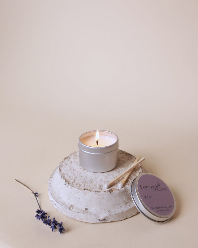 A lit natural soy candle tin from Love to b sits on a painted pot with some lavender next to it.