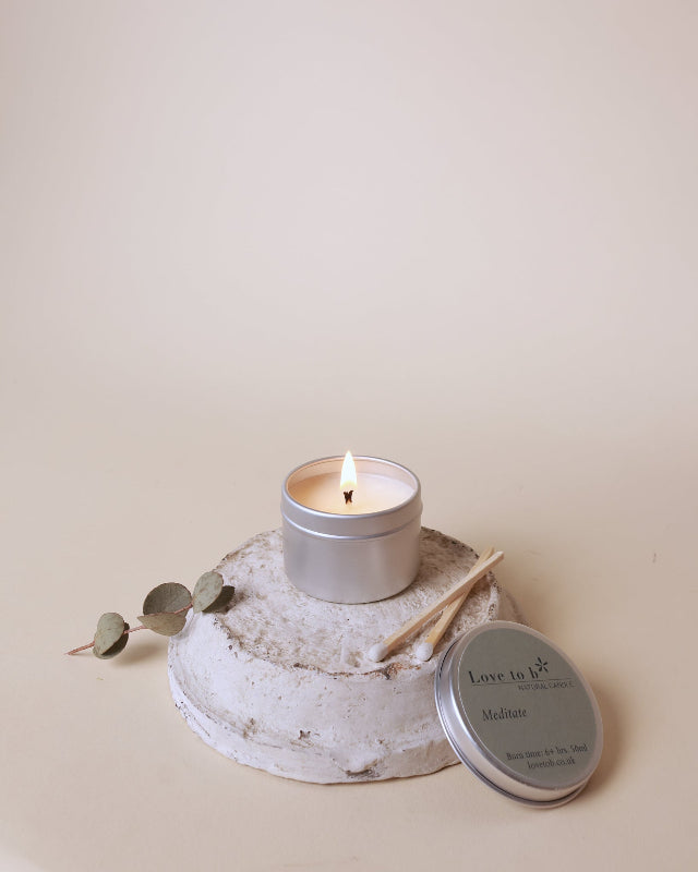 A lit silver tin natural candle sits on a round textured stone pedestal, with a wooden matchstick, a small eucalyptus sprig, and the matching silver lid placed.