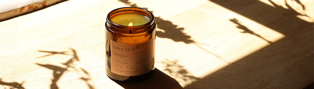 An amber glass jar natural soy candle from Love to b sits on a light wooden surface in direct sunlight, casting a shadow with leafy silhouettes.