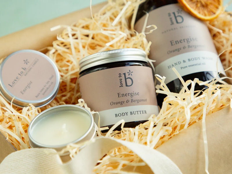 A gift set of all natural face, body and hand products from Love to b, including an Energise Orange & Bergamot body butter jar, a matching hand and body wash bottle, and a candle, arranged in a box filled with shredded paper.